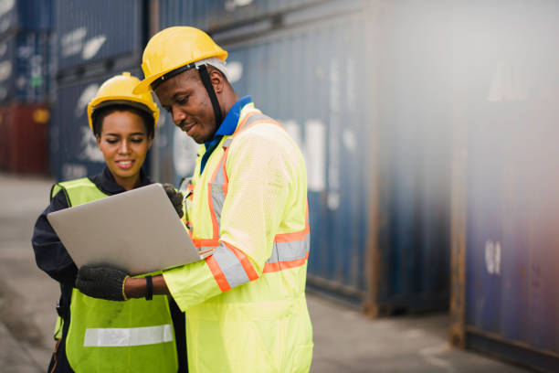 Young African american men and woman worker Check and control loading freight Containers by use computer laptop at commercial shipping dock felling happy. Cargo freight ship import export concept